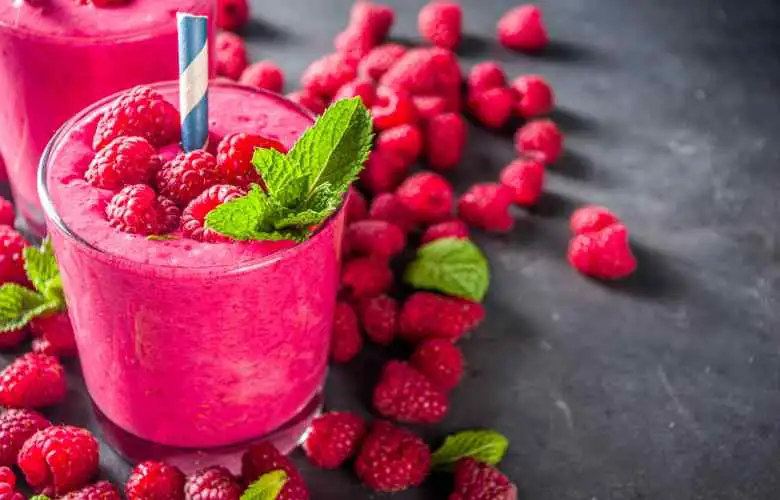 Two glasses of raspberry smoothie, also known as raspberry milkshake, surrounded by fresh raspberries.