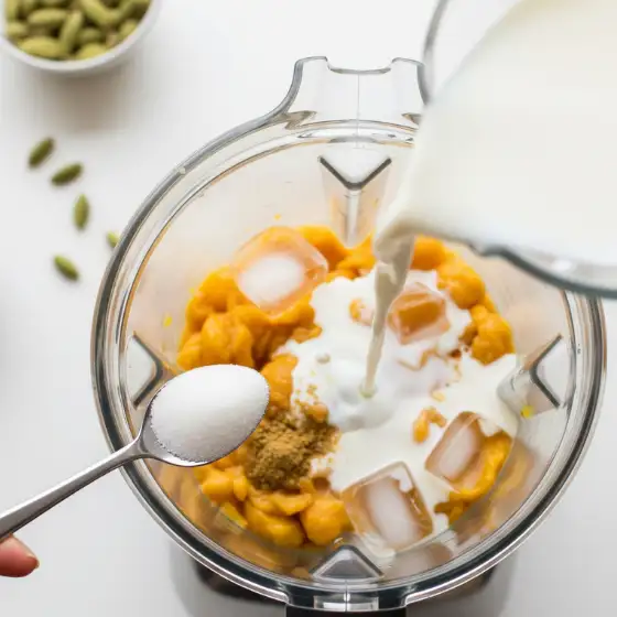 A blender filled with ingredients for a Chikoo Milkshake, including pumpkin puree, ice, and spices, as milk is poured in, with a spoon of sugar held above.