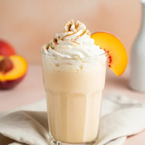 A creamy Peach Milkshake topped with whipped cream and cinnamon in a glass. A peach slice garnishes the rim. Soft, pastel background tones.
